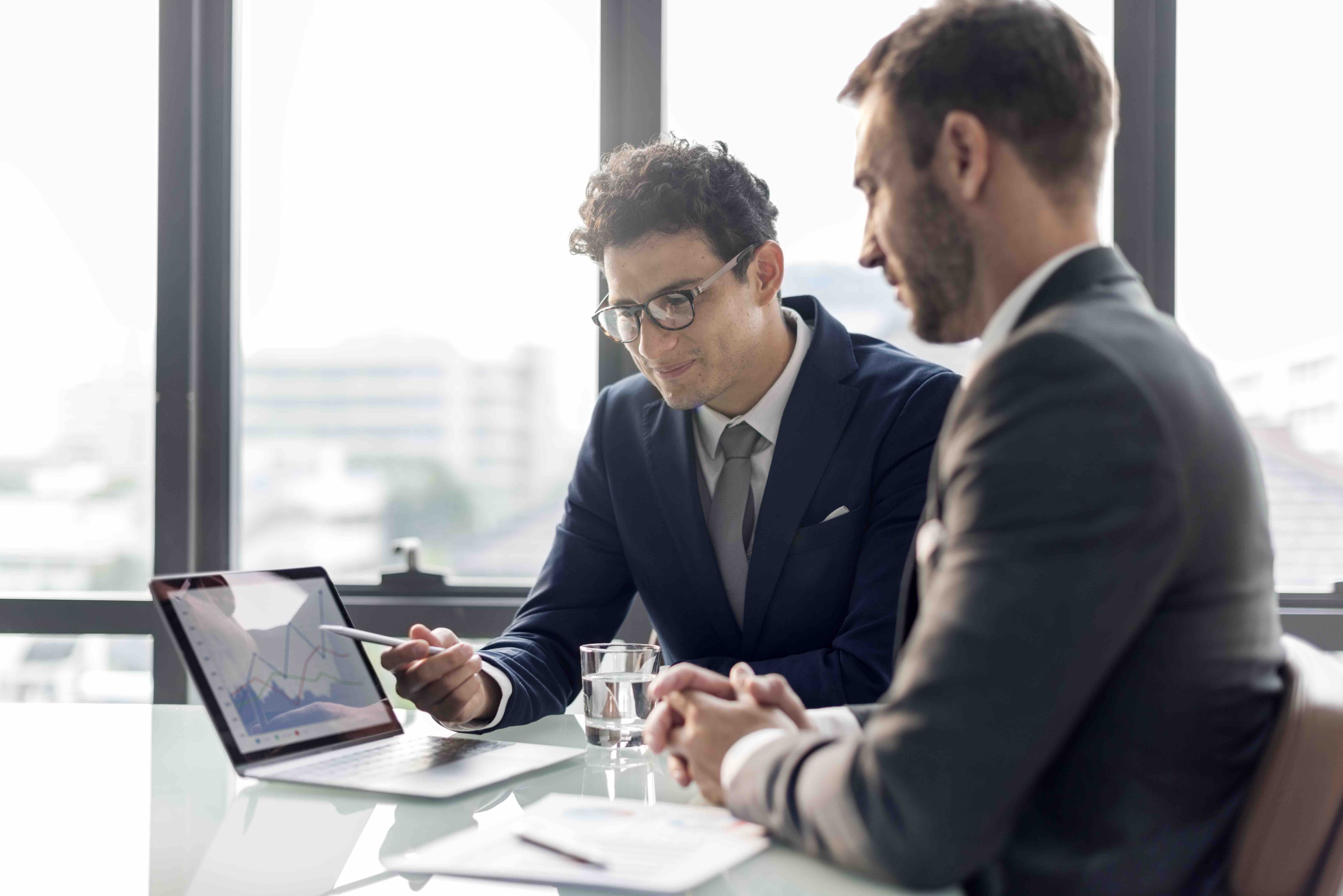 Two businessmen discussing charts on a laptop in a modern office.
